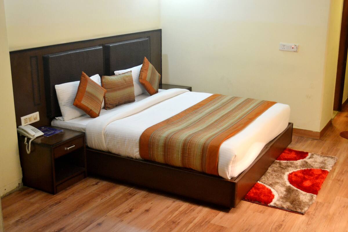 A neatly made bed with striped bedding, a wooden nightstand with a telephone, and a red and brown patterned rug on a wooden floor.