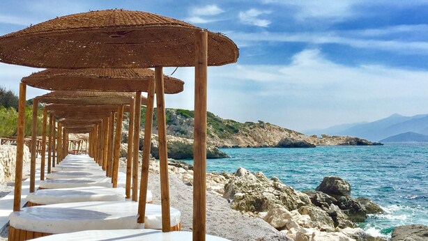 Une rangée de parasols de plage en bois, surmontés de coussins blancs, se dresse sur une plage de sable, offrant une vue sur la mer.