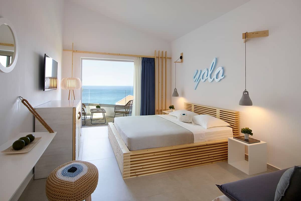 A modern bedroom with a bed, bedside tables, a small stool, a desk, and a view of the ocean.