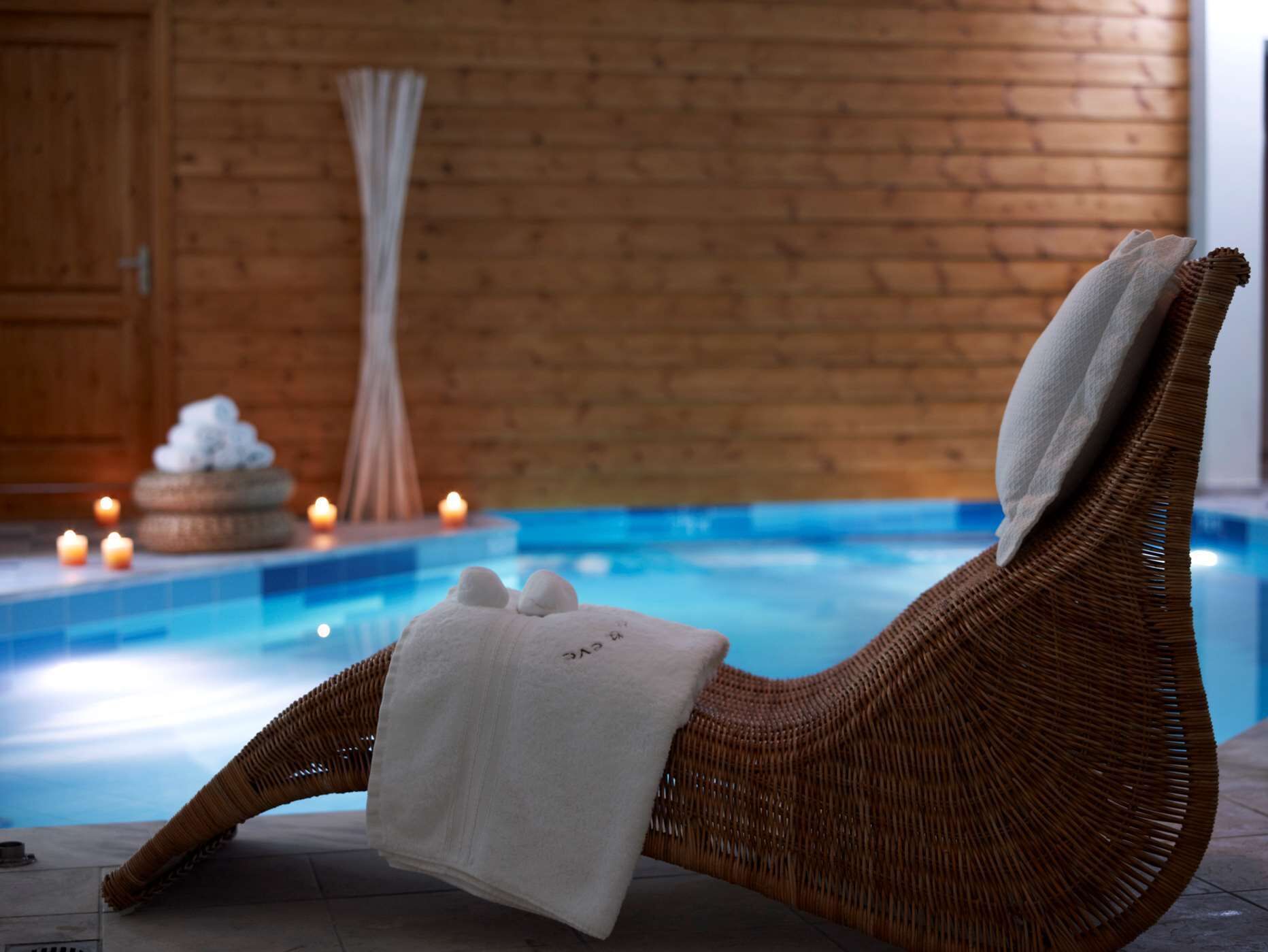 Una silla de mimbre para relajarse con una toalla, al lado de una piscina con velas y piedras al fondo.