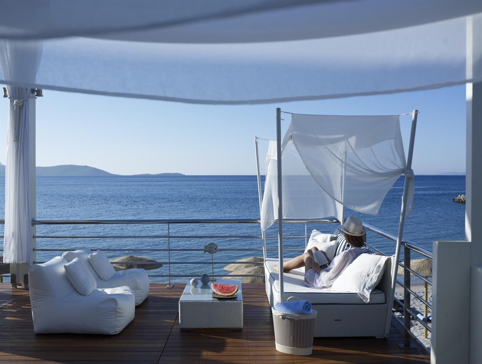 Una terraza tranquila con un toldo blanco, una silla reclinable con cojines, una mesita con sandía y un cubo de basura.