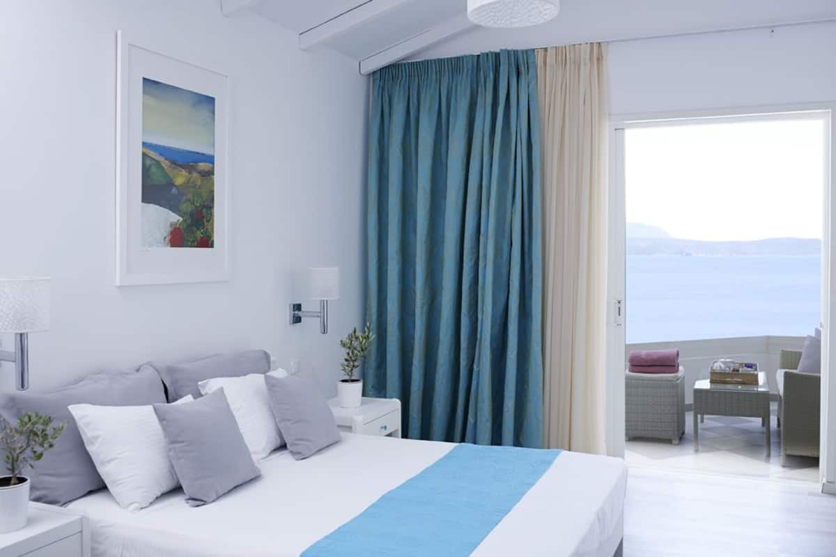 A modern hotel room with a bed, a nightstand, a lamp, a potted plant, and a view of the sea through a large window.