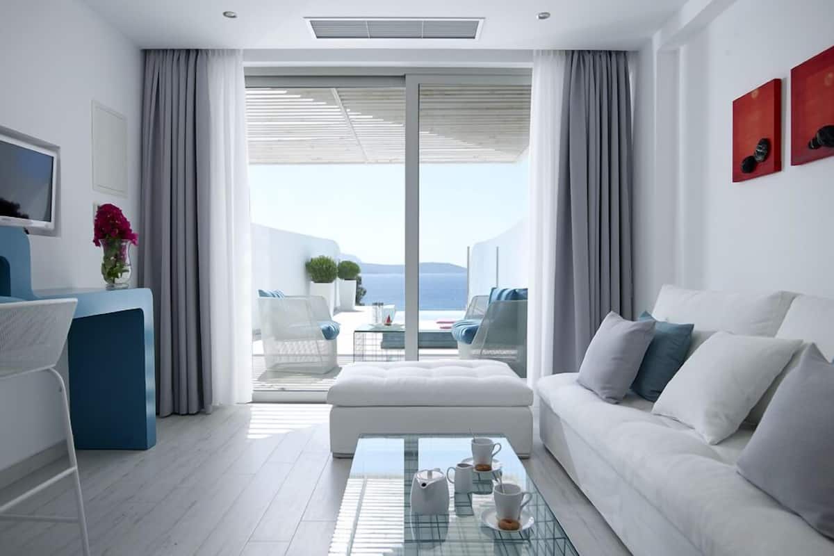A modern hotel room with a white sofa, a glass coffee table with cups, a blue desk, and a view of the sea through large windows.