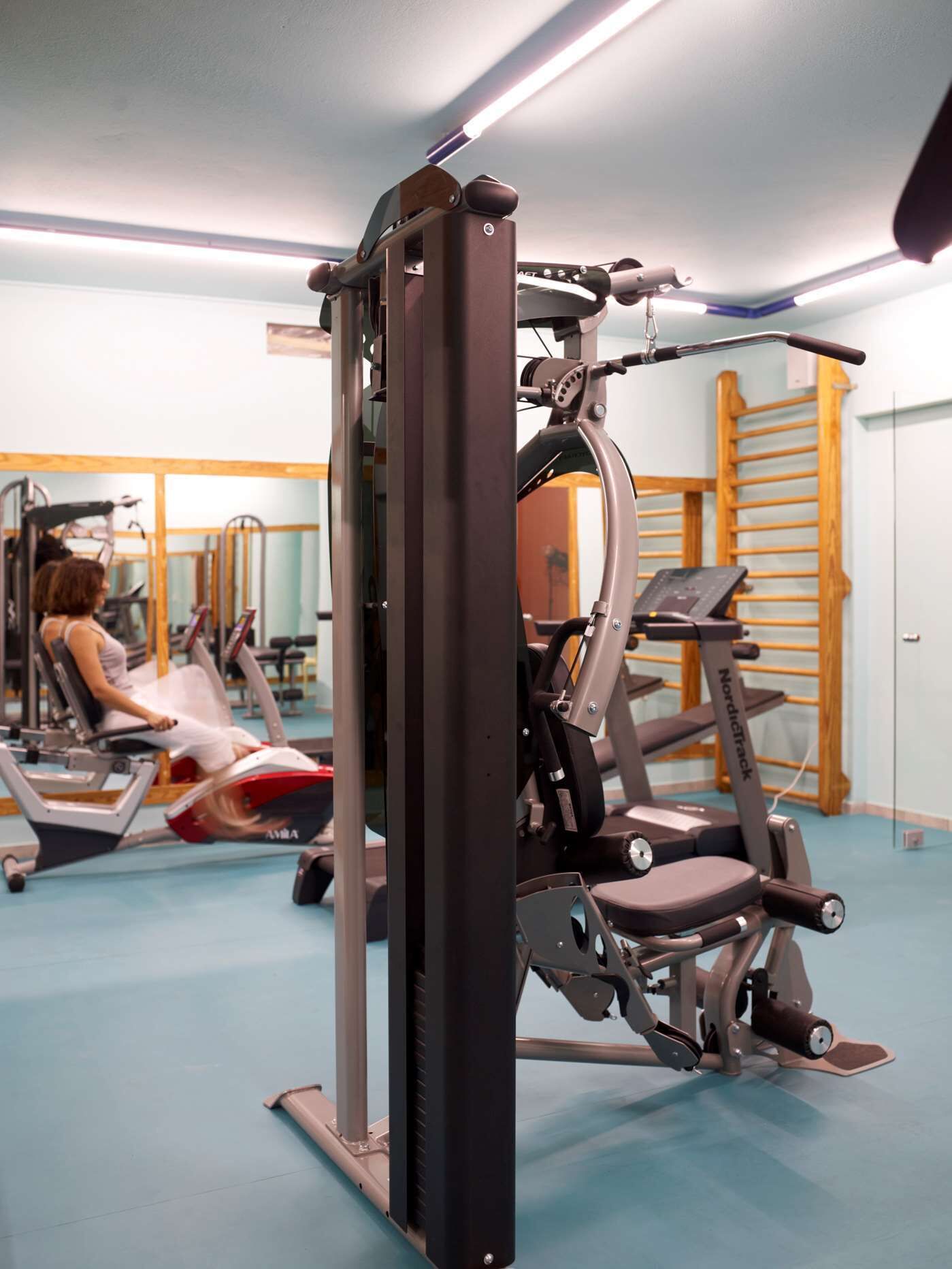 Ein Fitnessstudio mit Trainingsgeräten, Laufband und einer Person, die ein Gerät benutzt.