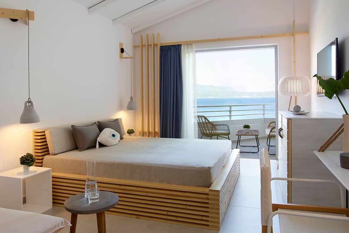 A modern hotel room with a large bed, a small table, a desk, and a balcony with a view of the sea.