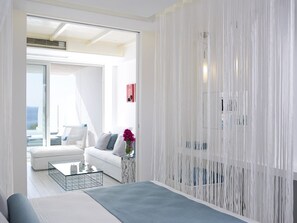 A modern hotel room with a large bed, a white sofa, a coffee table, and a view of the sea through a glass door. - Proteas Blu Resort - Adults Only (Samos)