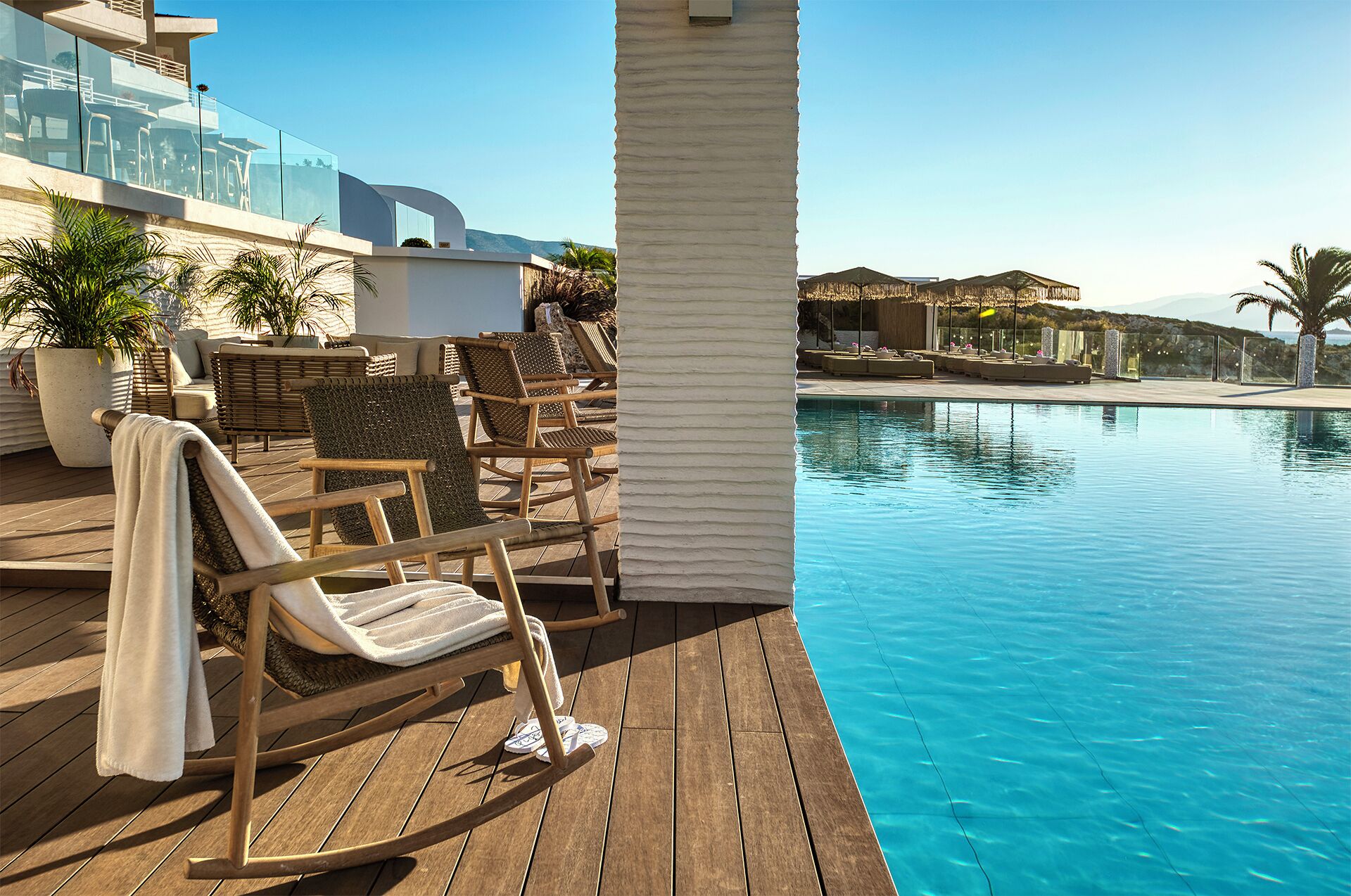 A poolside area with wooden deck chairs, a glass balustrade, and a view of a mountain range.