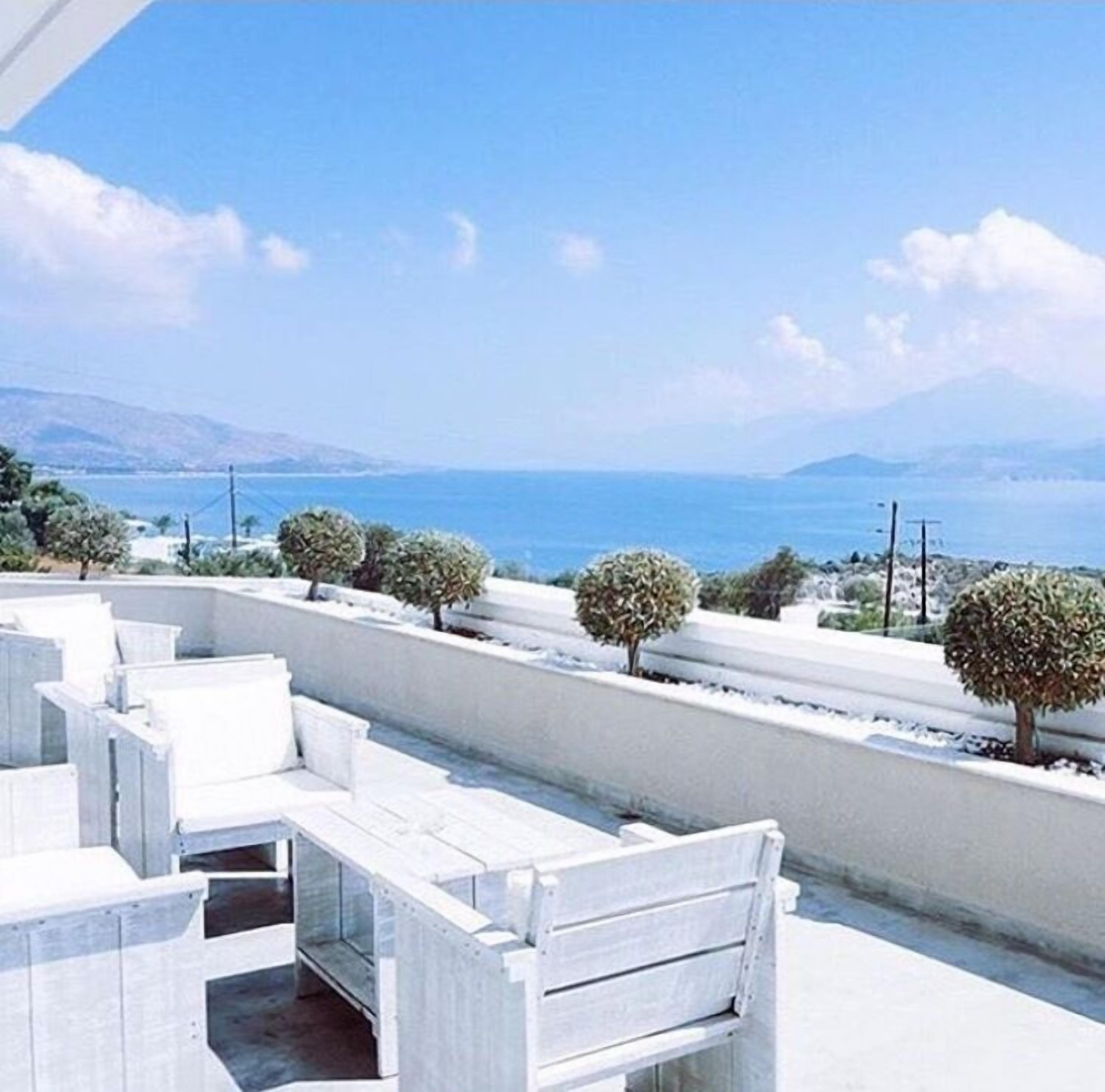 Un balcon avec des meubles blancs, une vue sur la mer et un paysage montagneux en arrière-plan.