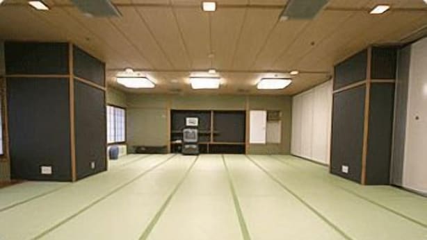 Una habitación con suelo de tatami, techo bajo con luces fluorescentes y una puerta corredera en un lateral.