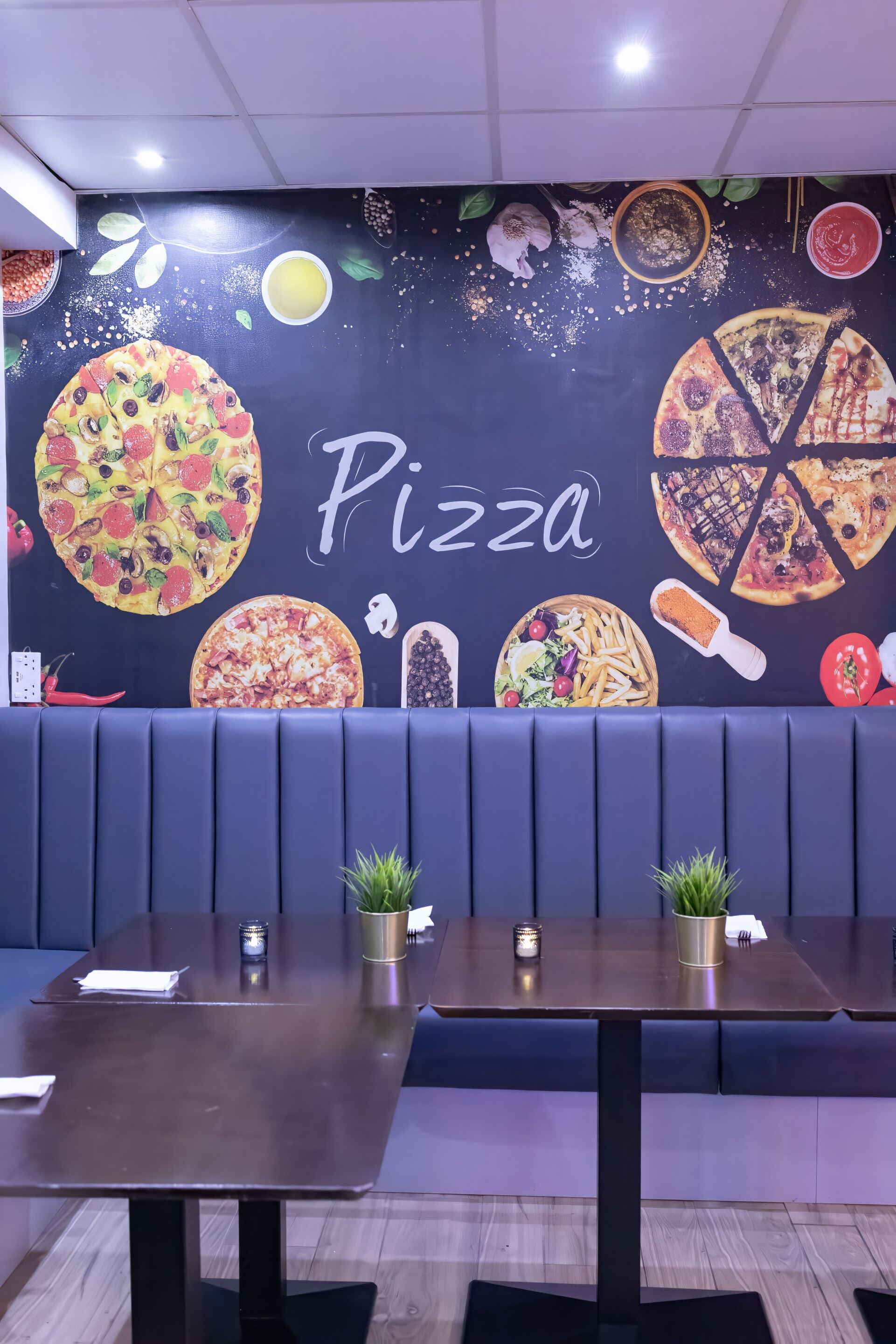 A restaurant interior with a wall mural of various pizzas, a seating area with booths, and a dark wood table.