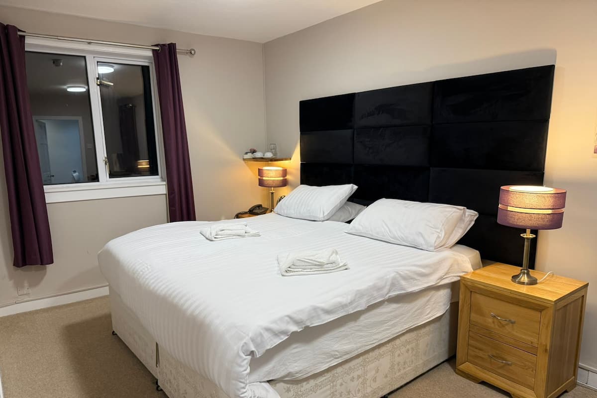Cama bem arrumada com lençóis brancos, luminária de cabeceira e mesa de cabeceira de madeira com gavetas.