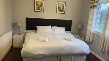 Una cama bien hecha con almohadas, una lámpara de noche, un cuadro en la pared y una ventana con cortinas.