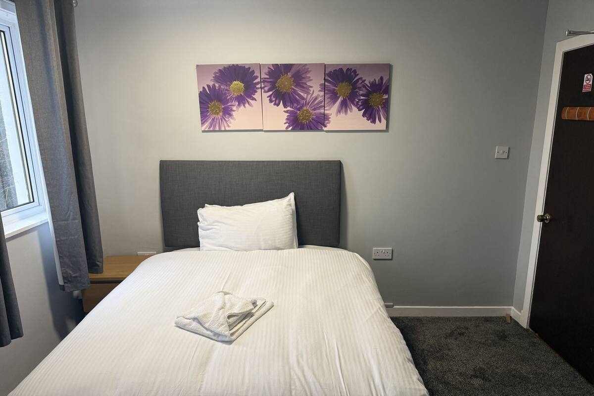 Cama bem arrumada com edredom branco e travesseiros, cabeceira cinza, quadro na parede com flores roxas e uma porta à direita.