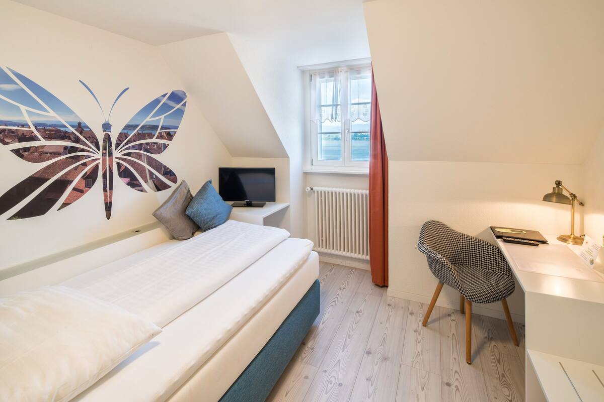 Un dormitorio con cama, escritorio, silla, televisión y ventana con vista a edificios.