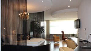 Una habitación de hotel moderna con una cama curvada, un candelabro, un televisor de pantalla plana y un comedor con sillas.