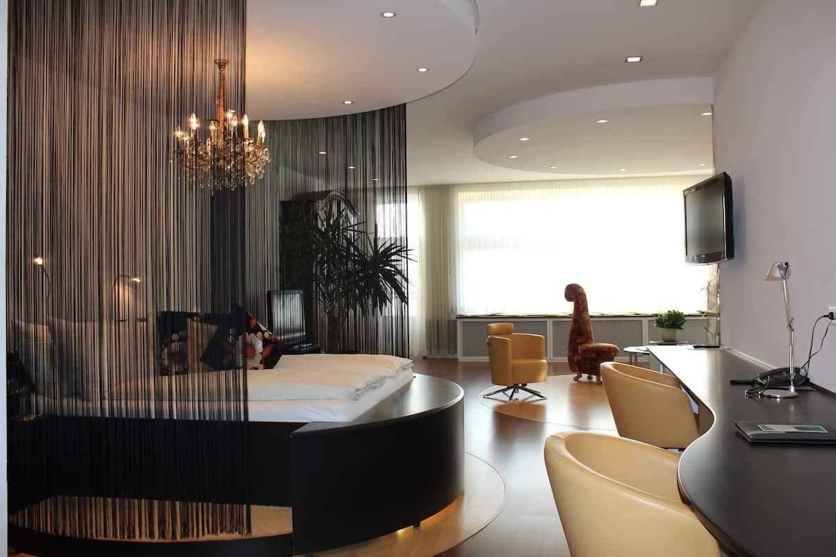 Una habitación de hotel moderna con una cama curvada, un candelabro, un televisor de pantalla plana y un comedor con sillas.