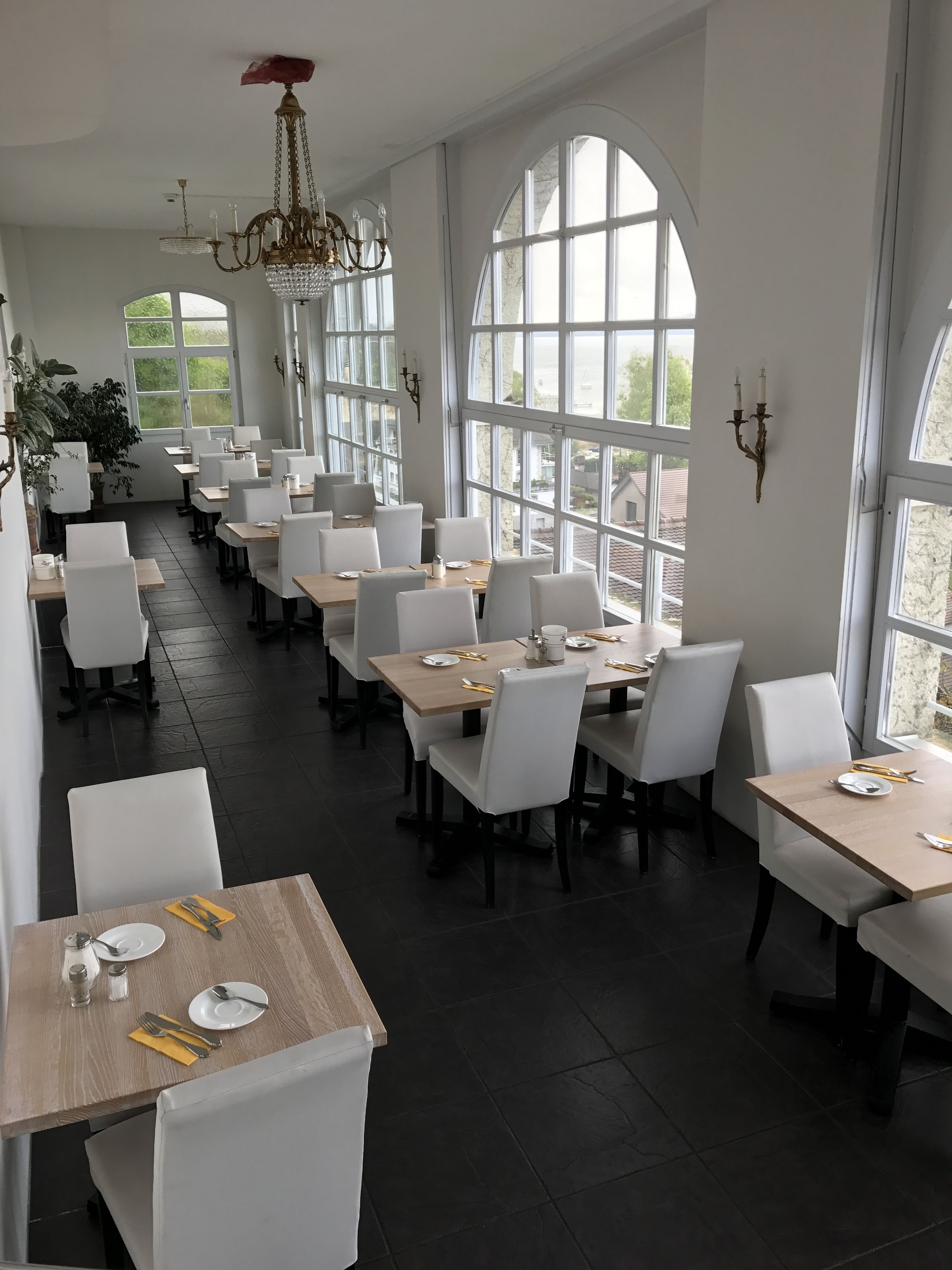 Un comedor amplio con techos altos, ventanas grandes y candelabros modernos. Las mesas están ordenadas con sillas blancas.