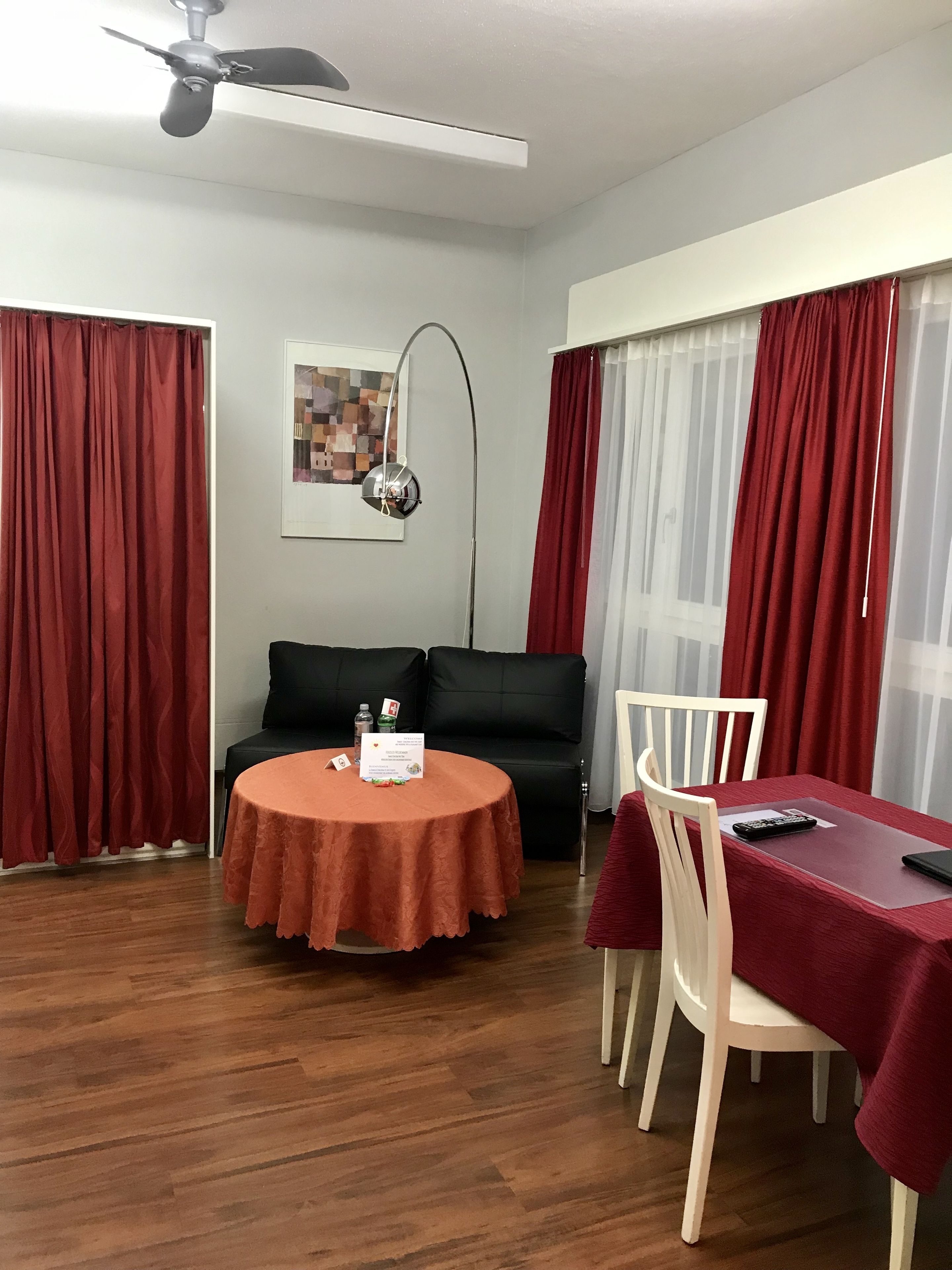 A room with a round table, a black sofa, a ceiling fan, and red curtains.