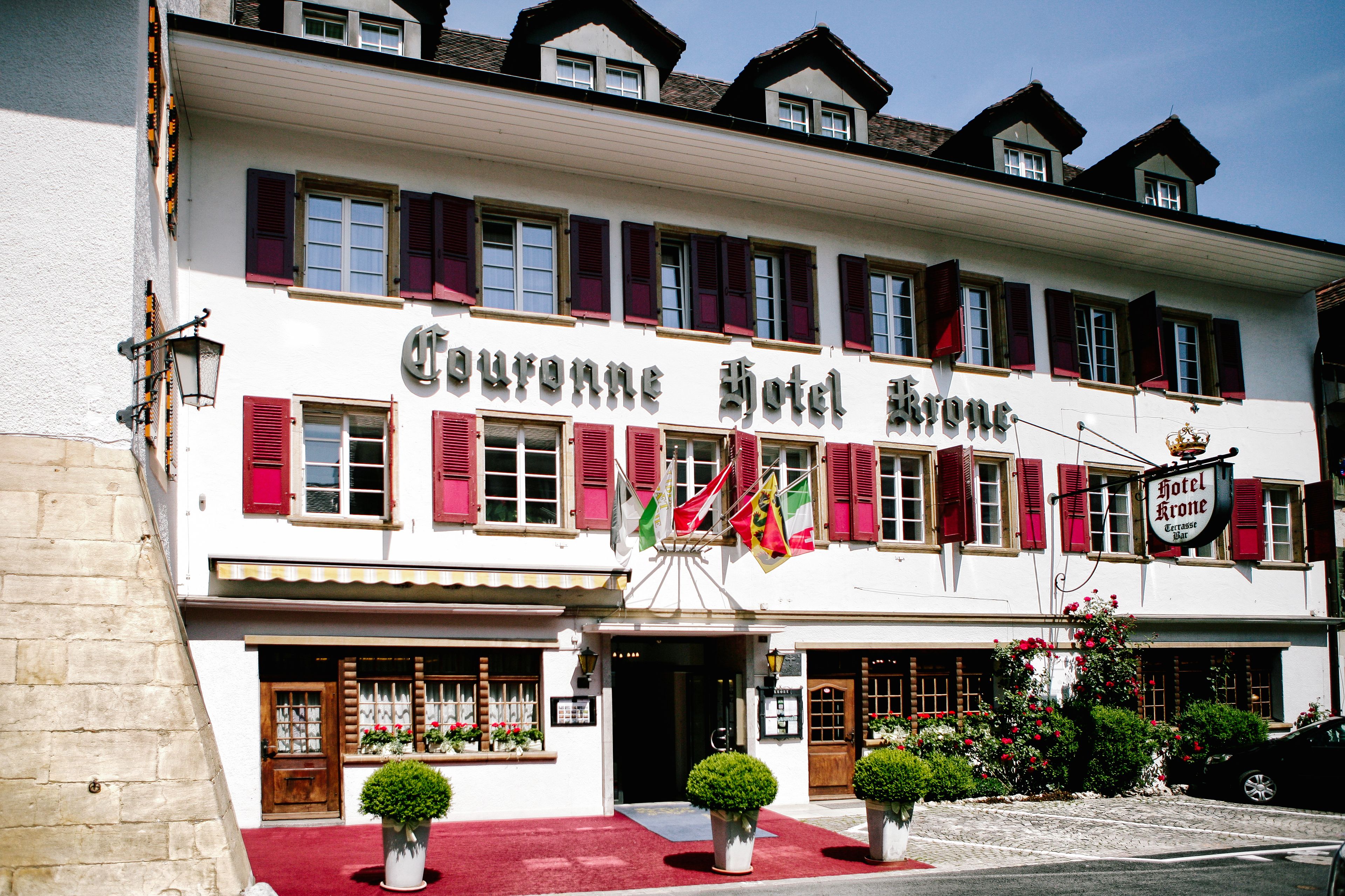 Un hôtel à la façade blanche, aux volets rouges, et dont l’enseigne indique « Couronne Hotel Krone ».