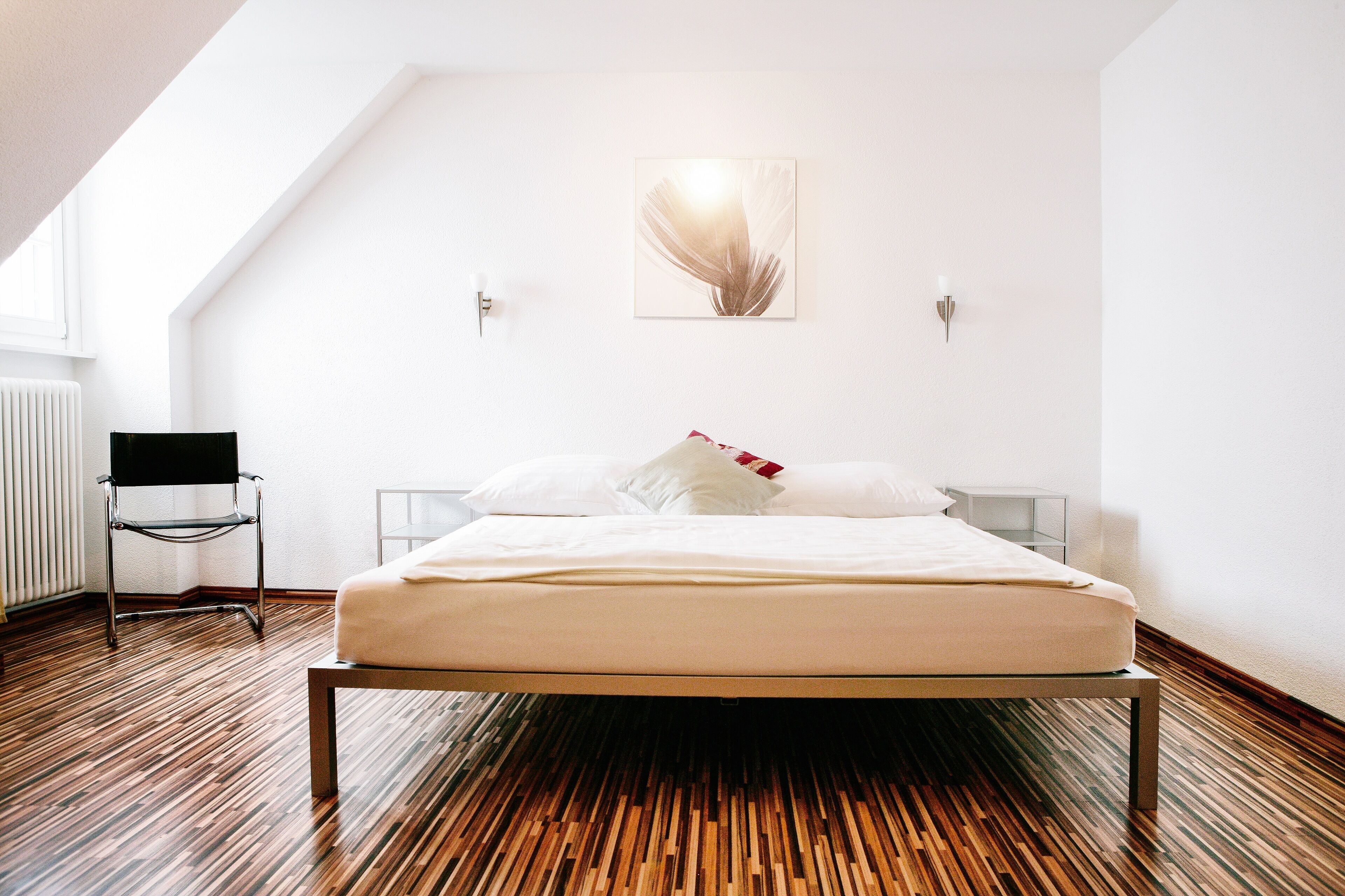 Camera da letto moderna con soffitto spiovente, un letto con lenzuola bianche, una sedia nera e un punto luce a parete.