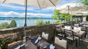 Un área de comedor al aire libre con mesas y sillas, grandes sombrillas y una vista panorámica de un lago y colinas.
