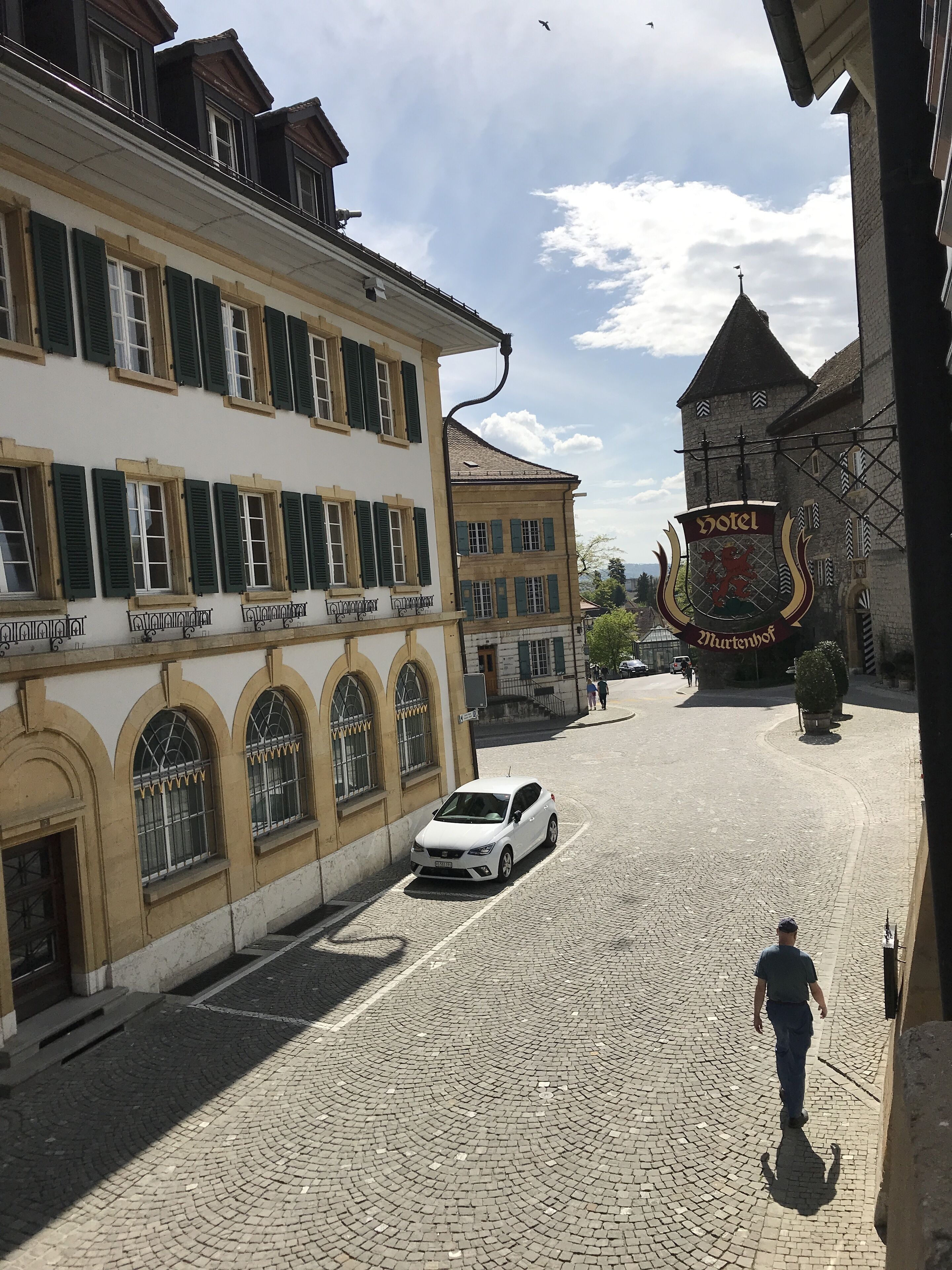 A cobblestone street with a white car parked on the side, a man walking, and a building with a sign that reads "Hotel Thurnenhof".