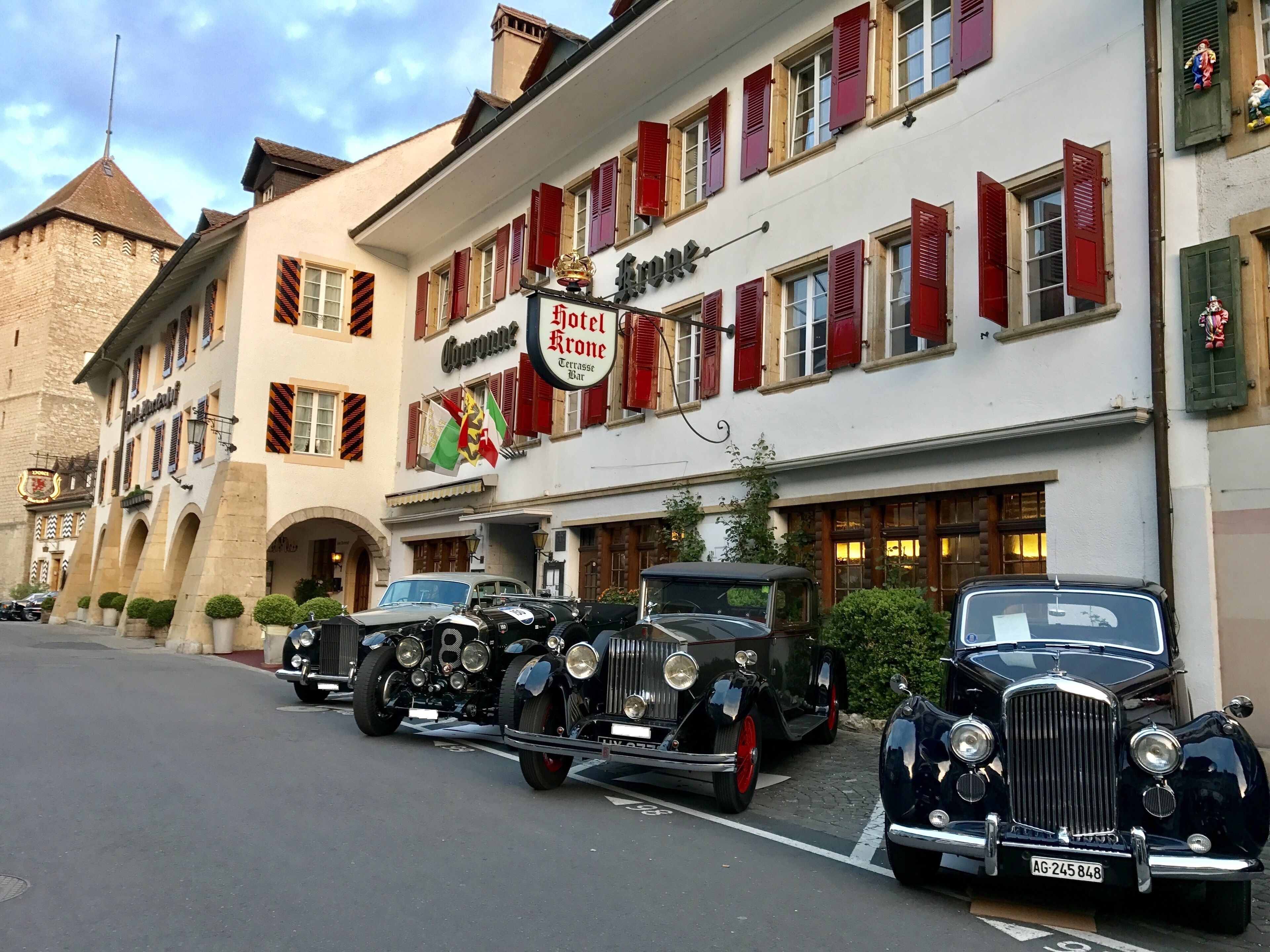 Un hôtel avec des volets rouges, une enseigne et des voitures anciennes garées devant.