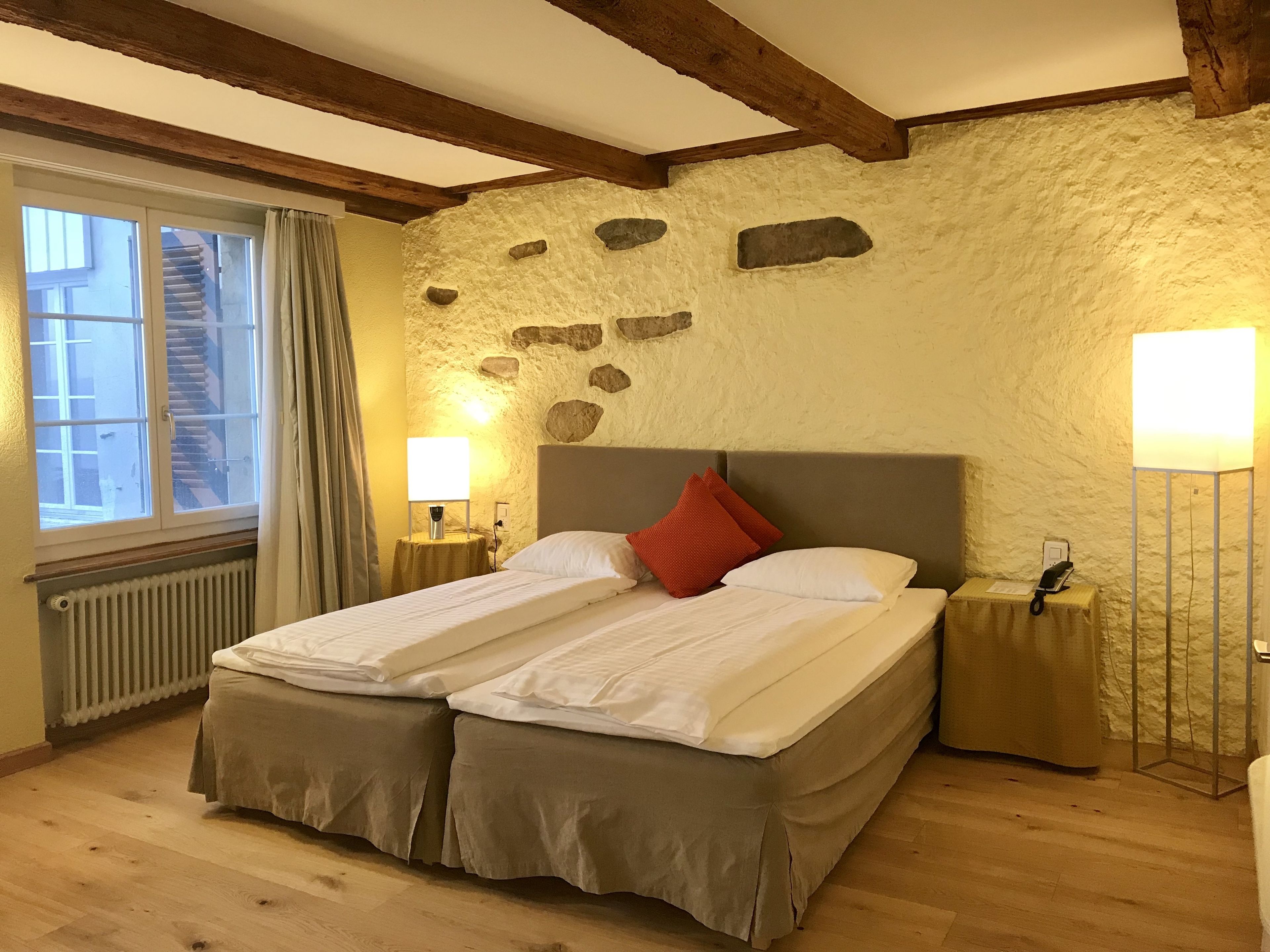 A double bed with a grey headboard, two bedside tables with lamps, a stone wall, a window with curtains, and a radiator.