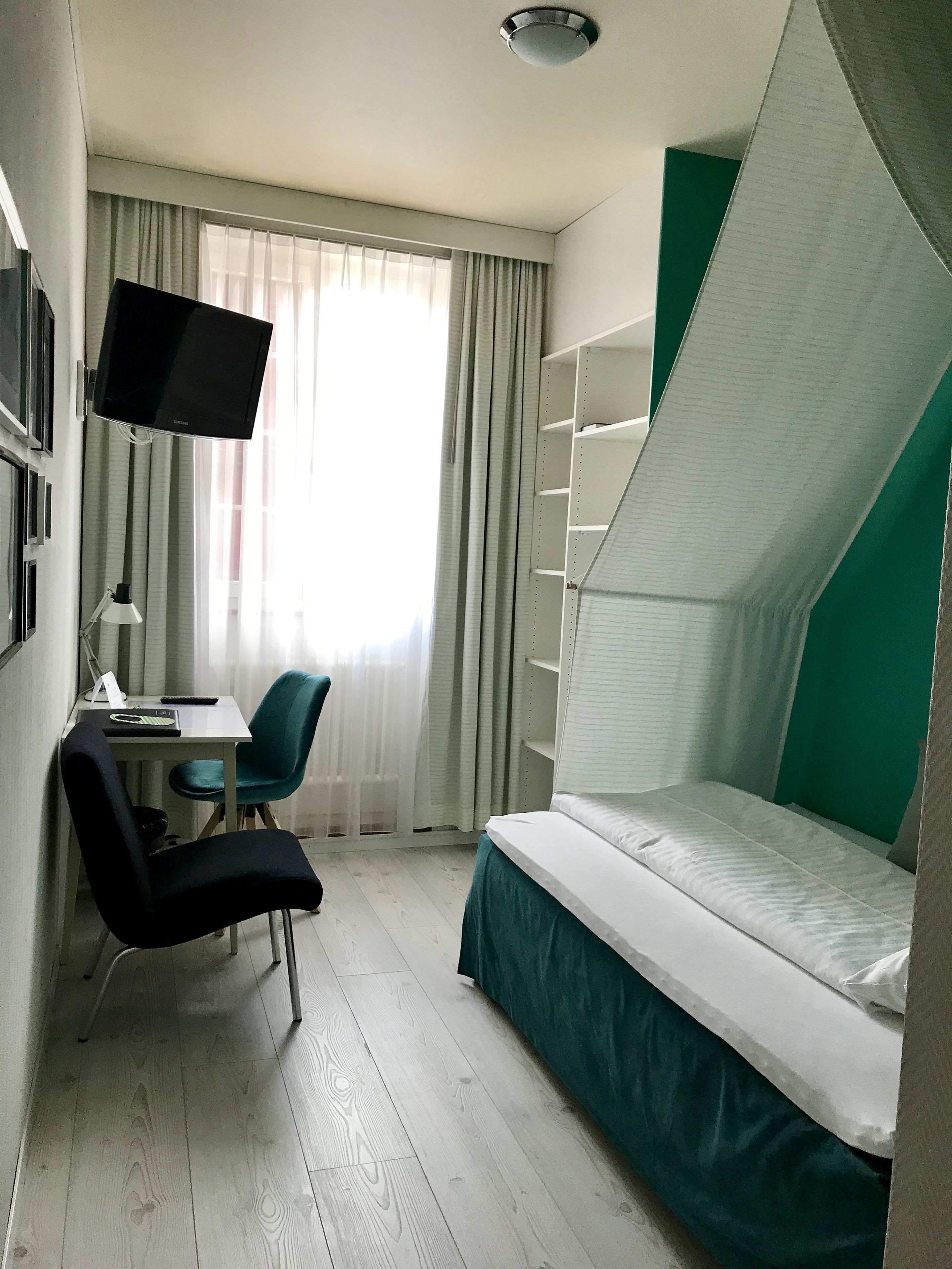 A compact hotel room with a single bed, a desk with a chair, a television mounted on the wall, and a window with curtains.