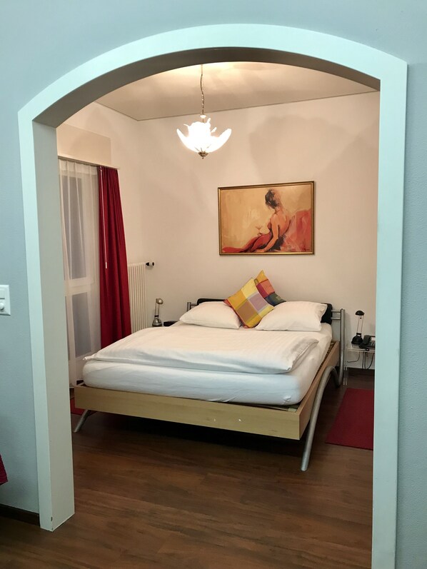 A bedroom with a bed, a painting, a nightstand, and a lamp.