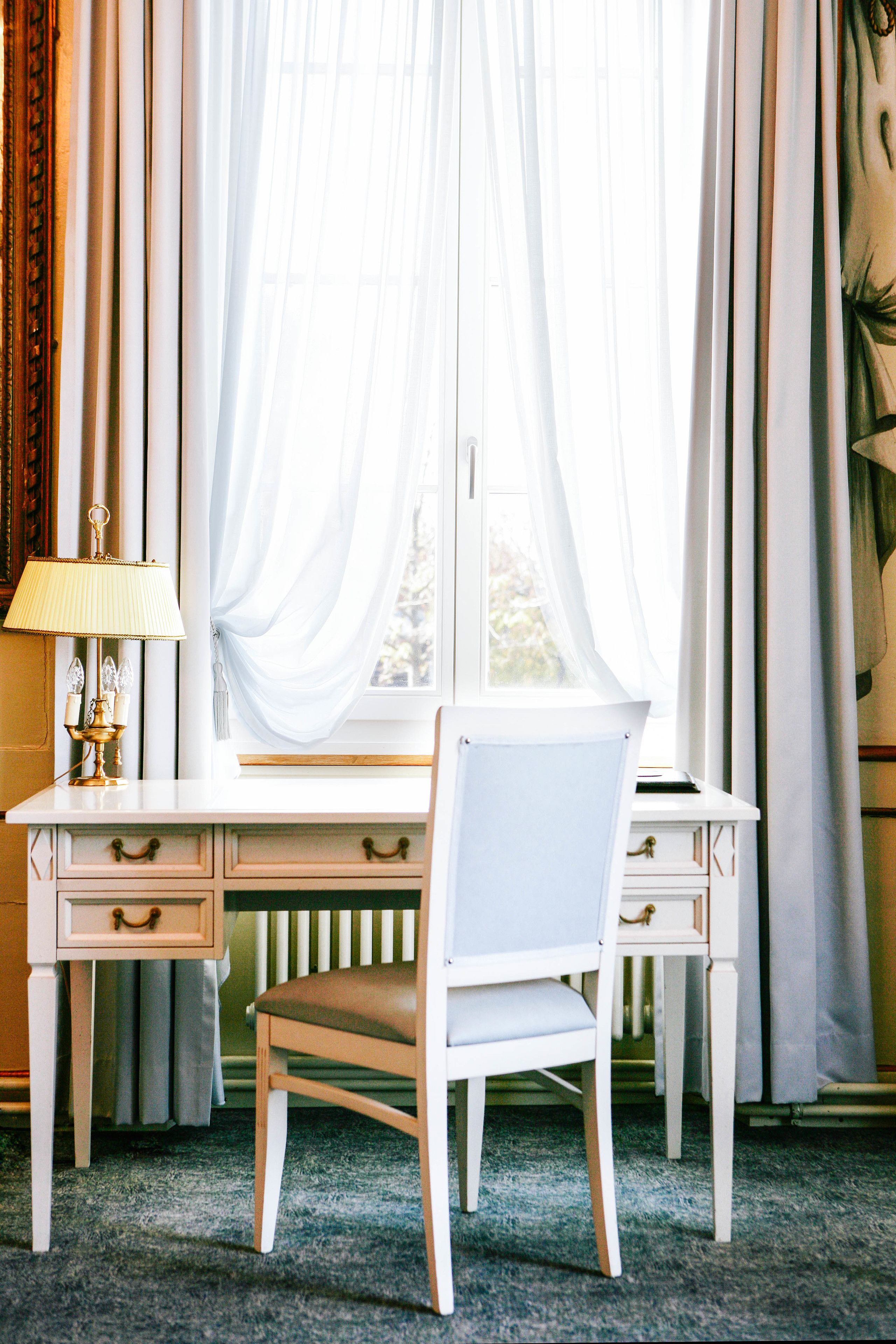 Ein Raum mit einem weißen Schreibtisch, einem Stuhl, einer Lampe und einem Fenster mit transparenten Vorhängen.
