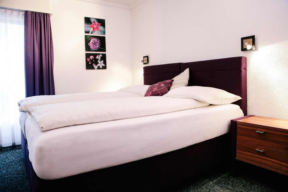 Una cama bien hecha con cabecera, una mesita de noche de madera con cajones, y un marco de pared con una obra de arte floral.