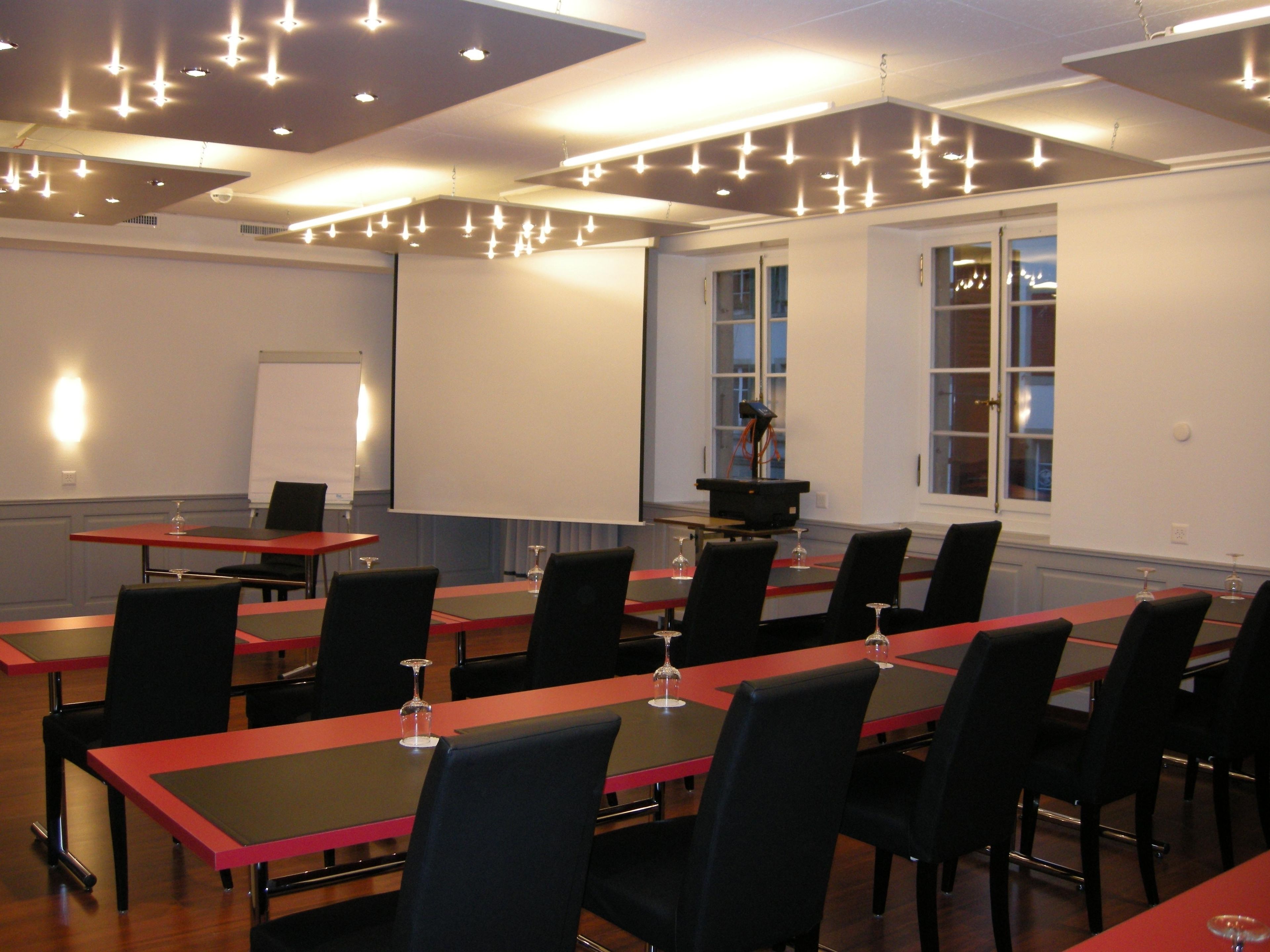 Ein Konferenzraum mit mehreren Tischen und Stühlen, einer Projektionsleinwand und einem Fenster mit Blick nach außen.
