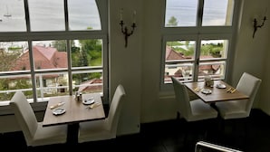 Comedor con dos mesas y sillas, ventanas arqueadas amplias con vista a un cuerpo de agua y edificios.