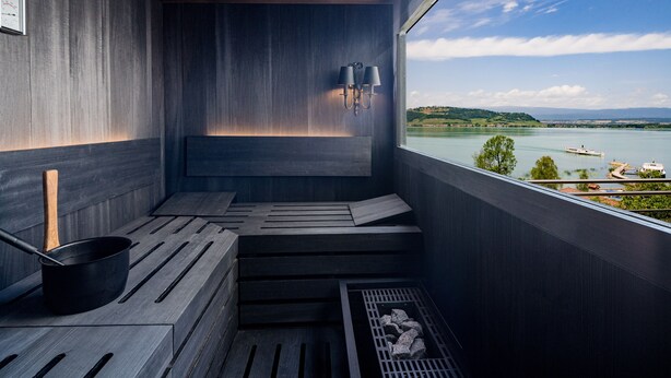 Una sauna con vistas a un lago y montañas, bancos de madera y una olla negra.