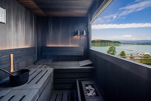 A sauna with a view of a lake and mountains, wooden benches, and a black pot.