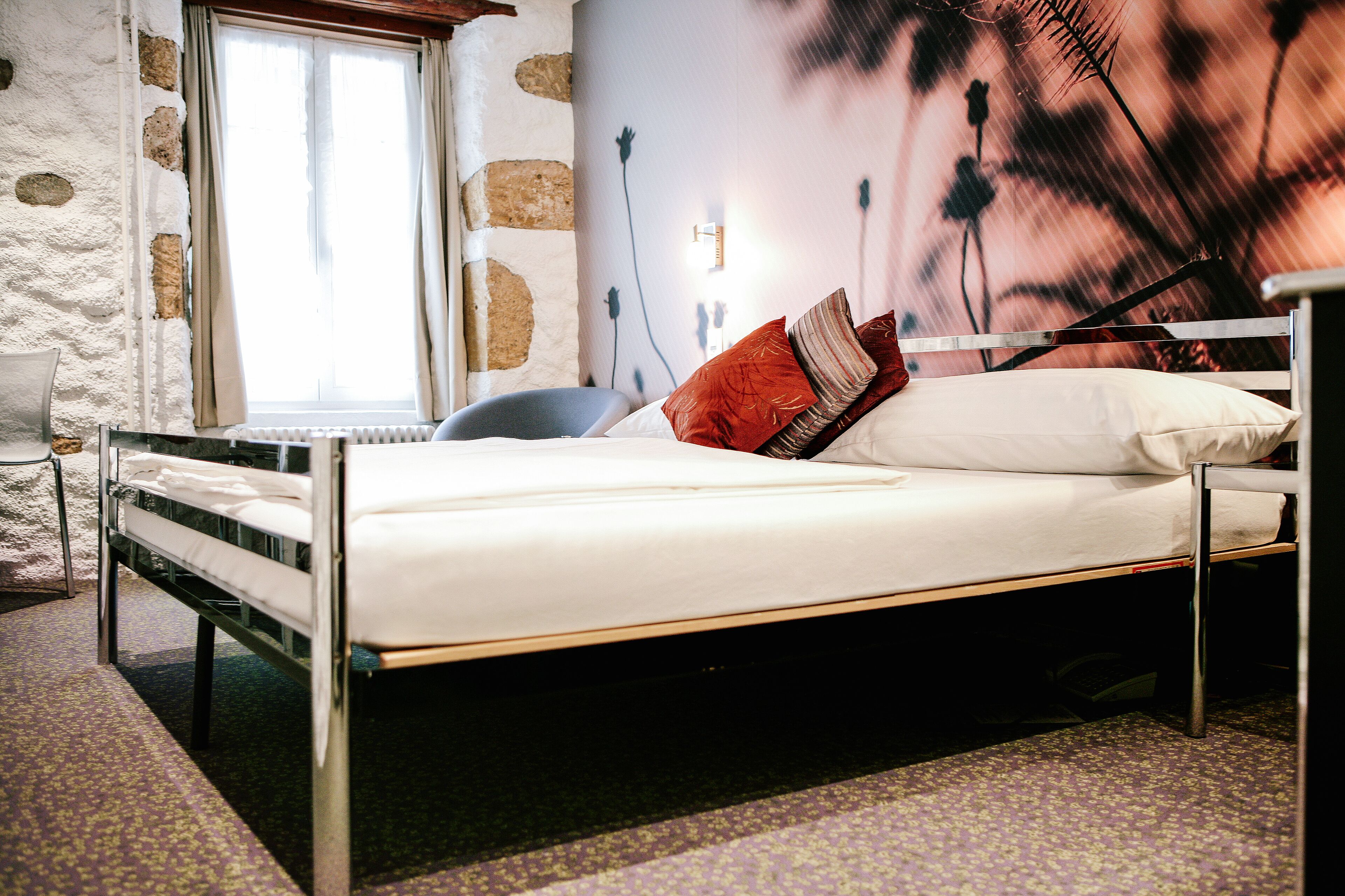 Ein Bett mit weißer Bettwäsche und einem Metallgestell, ein Stuhl, ein Heizkörper und eine Wand mit floralem Wandbild.