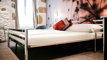Ein Bett mit weißer Bettwäsche und einem Metallgestell, ein Stuhl, ein Heizkörper und eine Wand mit floralem Wandbild.