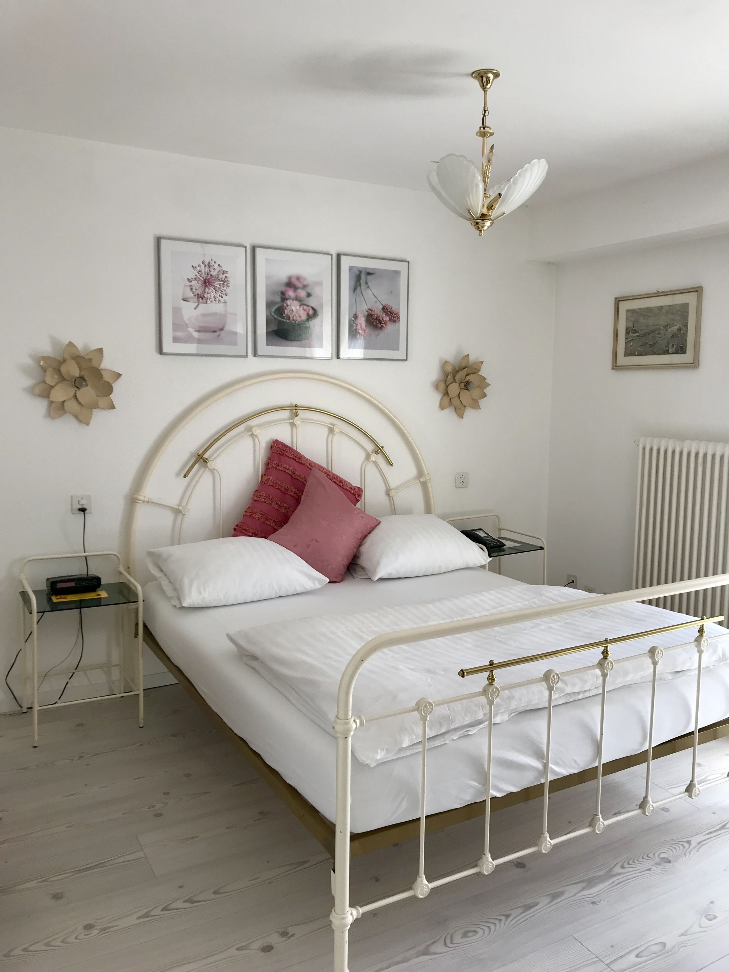 Ein ordentlich eingerichtetes Schlafzimmer mit einem weißen Metallbettgestell, einem Nachttisch mit Lampe und blumengeschmückten Wanddekorationen.
