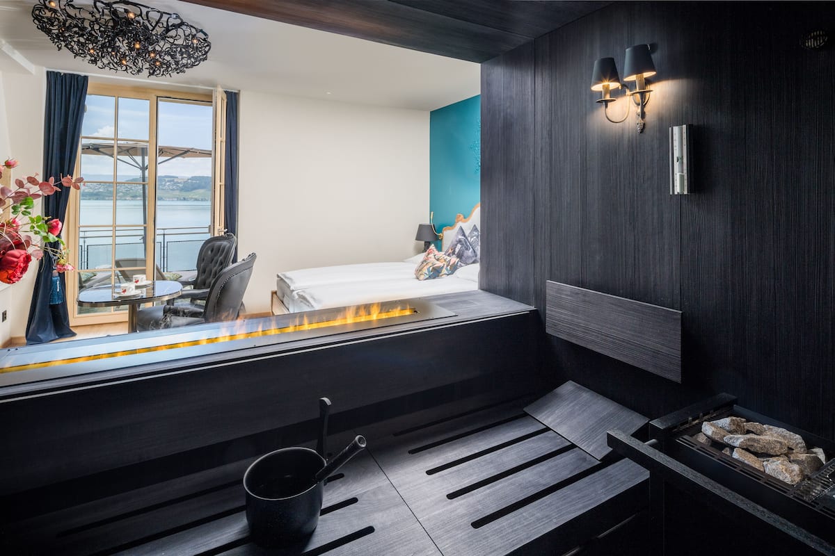 Una habitación de hotel moderna con un ventanal grande, una cama con chimenea, una mesita con sillas y vista al agua.