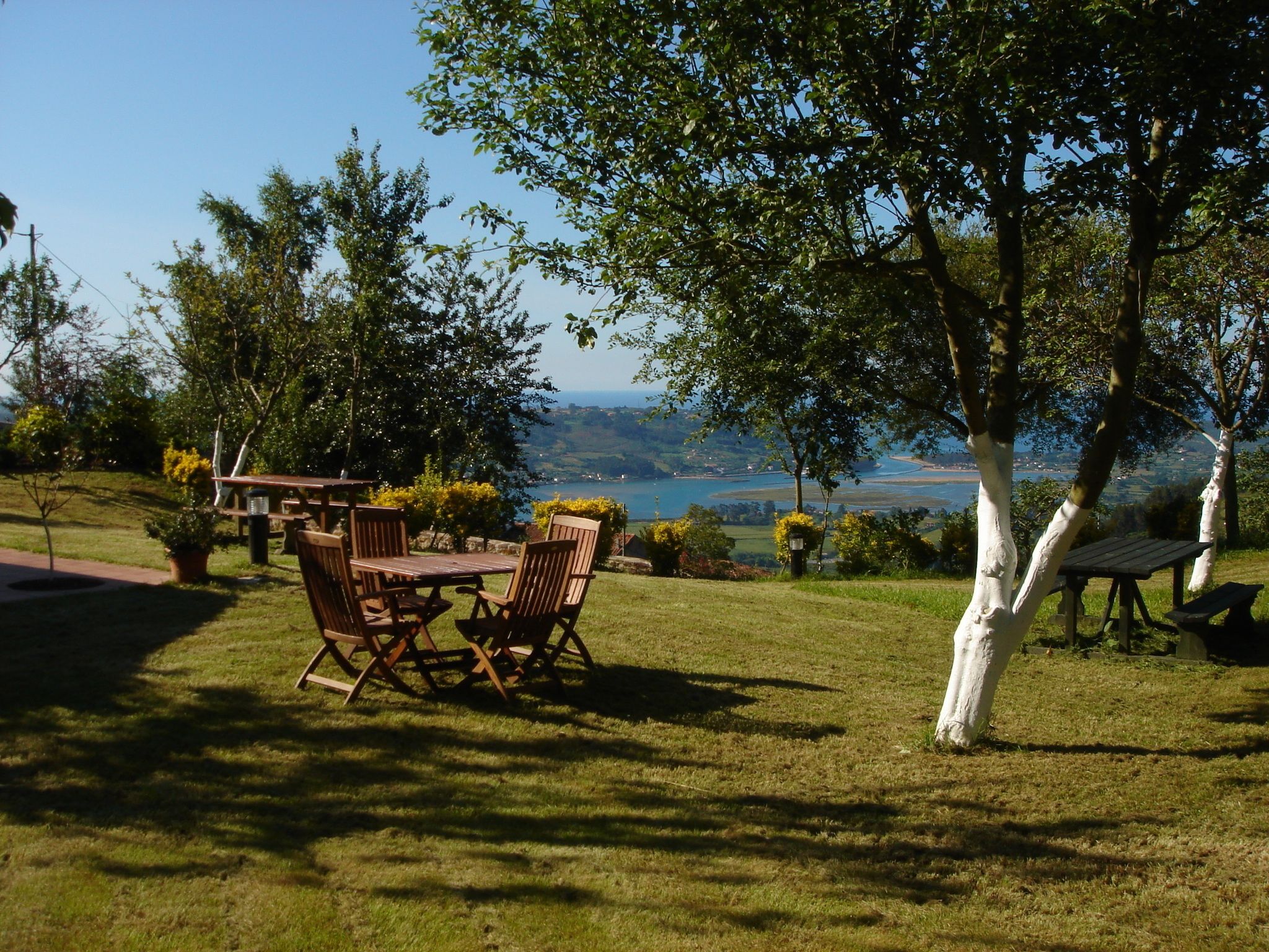 An outdoor seating area with a table and chairs, surrounded by trees and a view of a body of water and distant landscape.