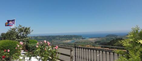 A balcony with a view of a coastal landscape, a flag, and a garden with pink flowers.