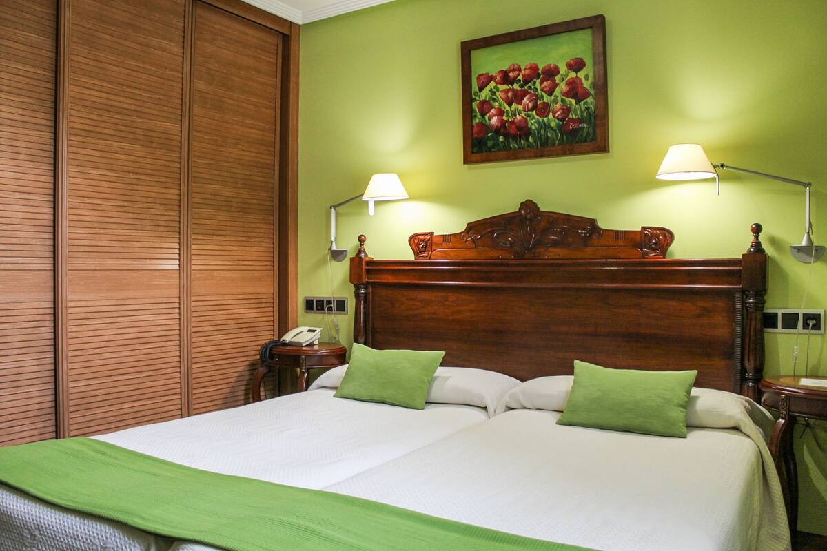 Una cama doble con almohadas verdes, cabecero de madera, dos lámparas de noche, un teléfono y un cuadro de flores rojas en la pared.