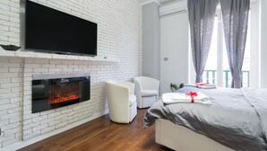 Een moderne hotelkamer met een witte baksteenmuur, een flatscreen televisie boven een open haard, een groot bed met grijze beddengoed, een witte fauteuil en een raam met grijze gordijnen.