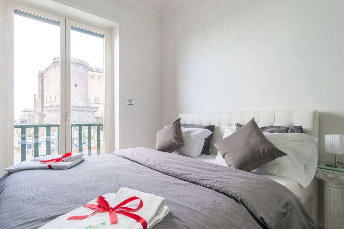 Een hotelkamer met een groot bed, grijze beddengoed en uitzicht op een kasteel door het raam.