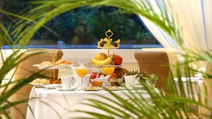 Una mesa con mantel blanco presenta una disposición escalonada de fruta y pastelería, una jarra de zumo de naranja y un arreglo de frutas tropicales.
