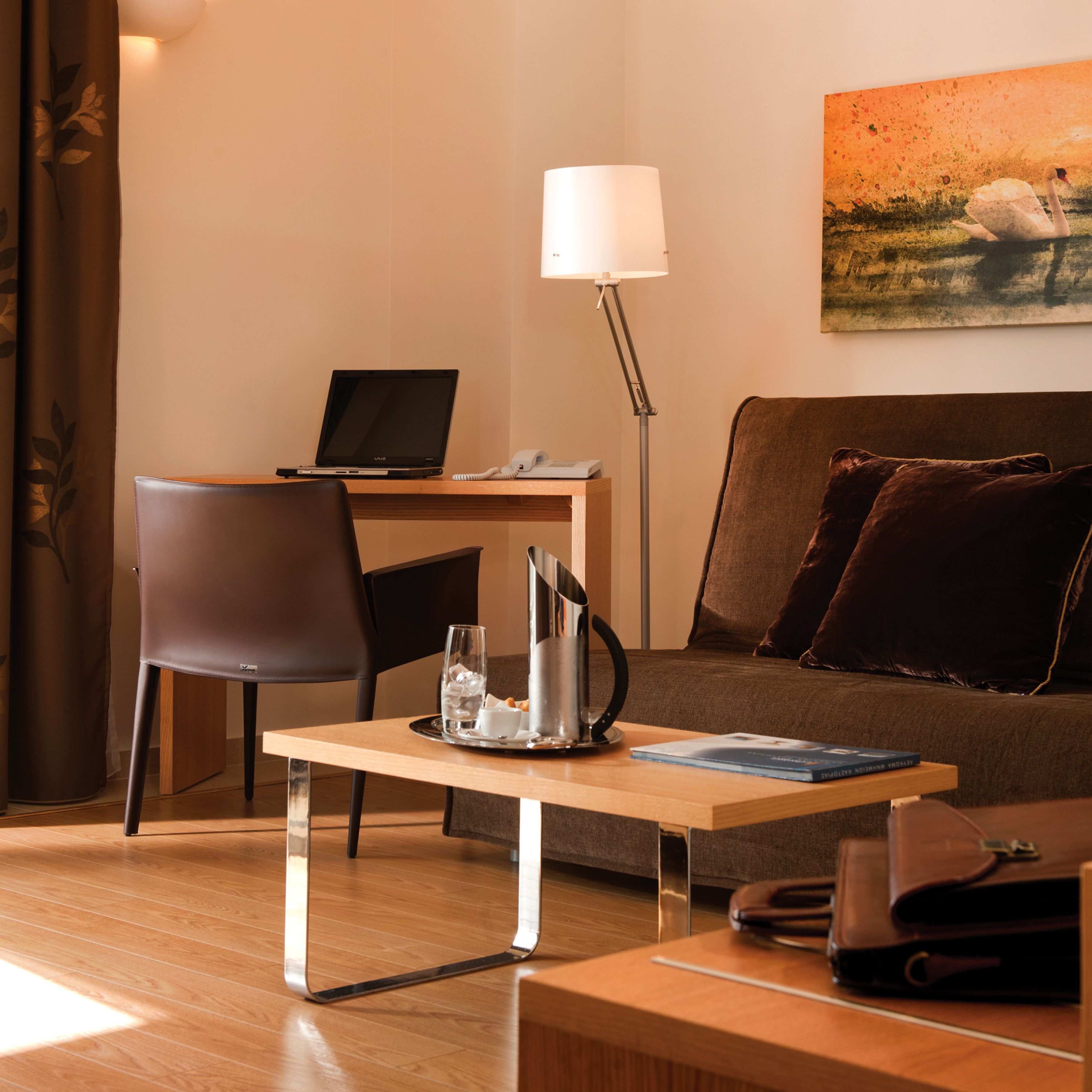 A modern hotel room with a wooden desk, a laptop, a lamp, a sofa with cushions, a coffee table, and a painting on the wall.