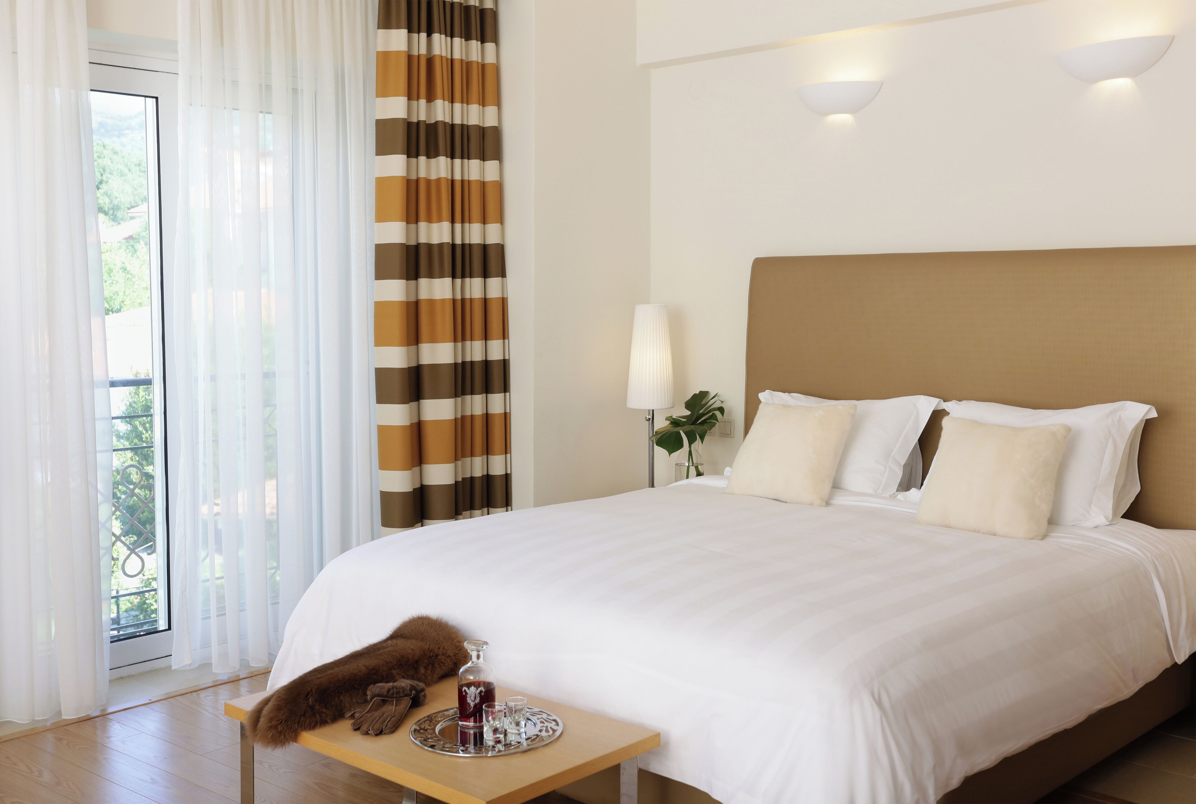 A hotel room with a large bed, a bedside table with a tray of drinks, and a view of greenery outside.