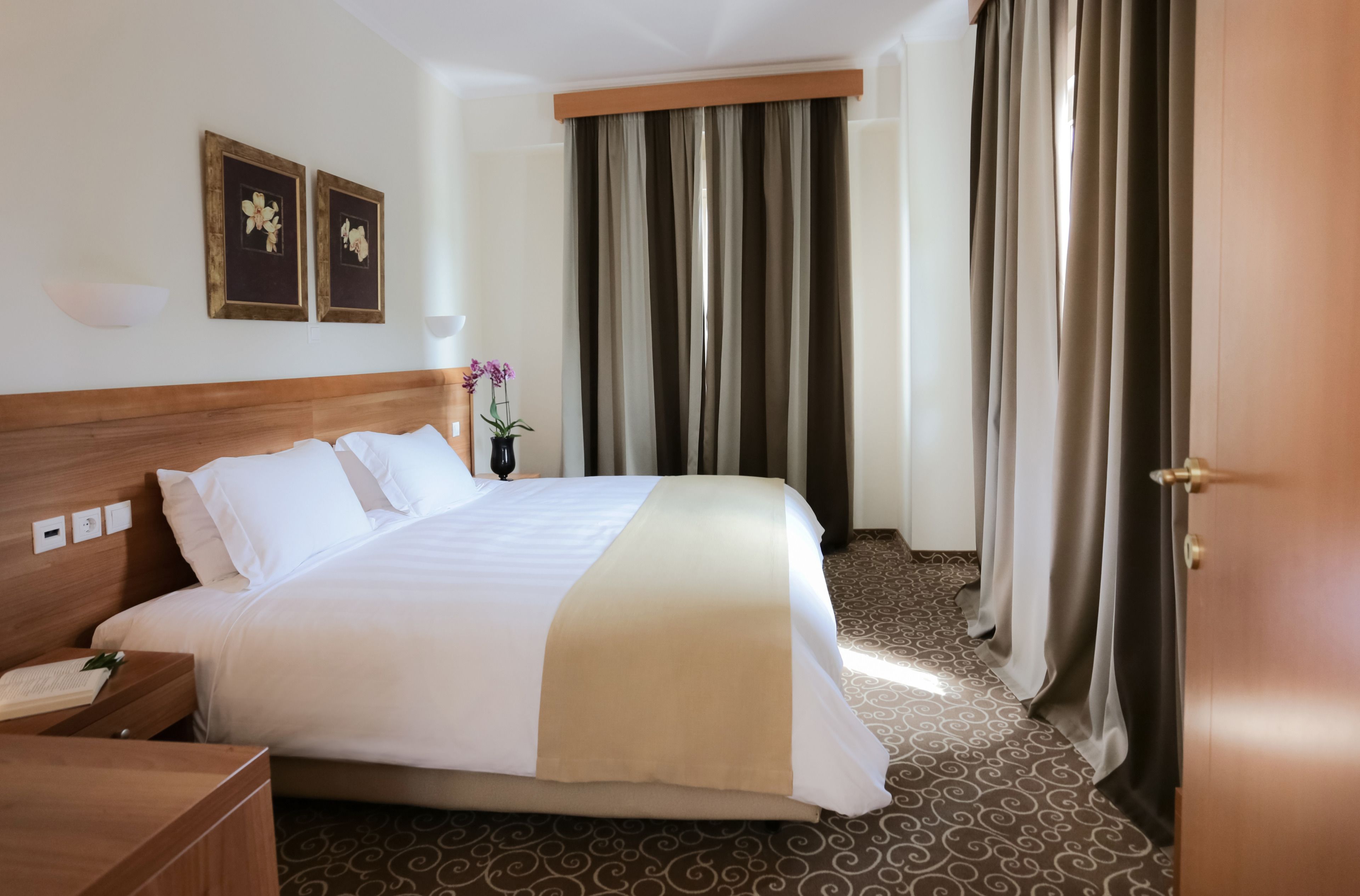 A hotel room with a large bed, a wooden headboard, two framed pictures on the wall, a nightstand, a lamp, and a window with curtains.