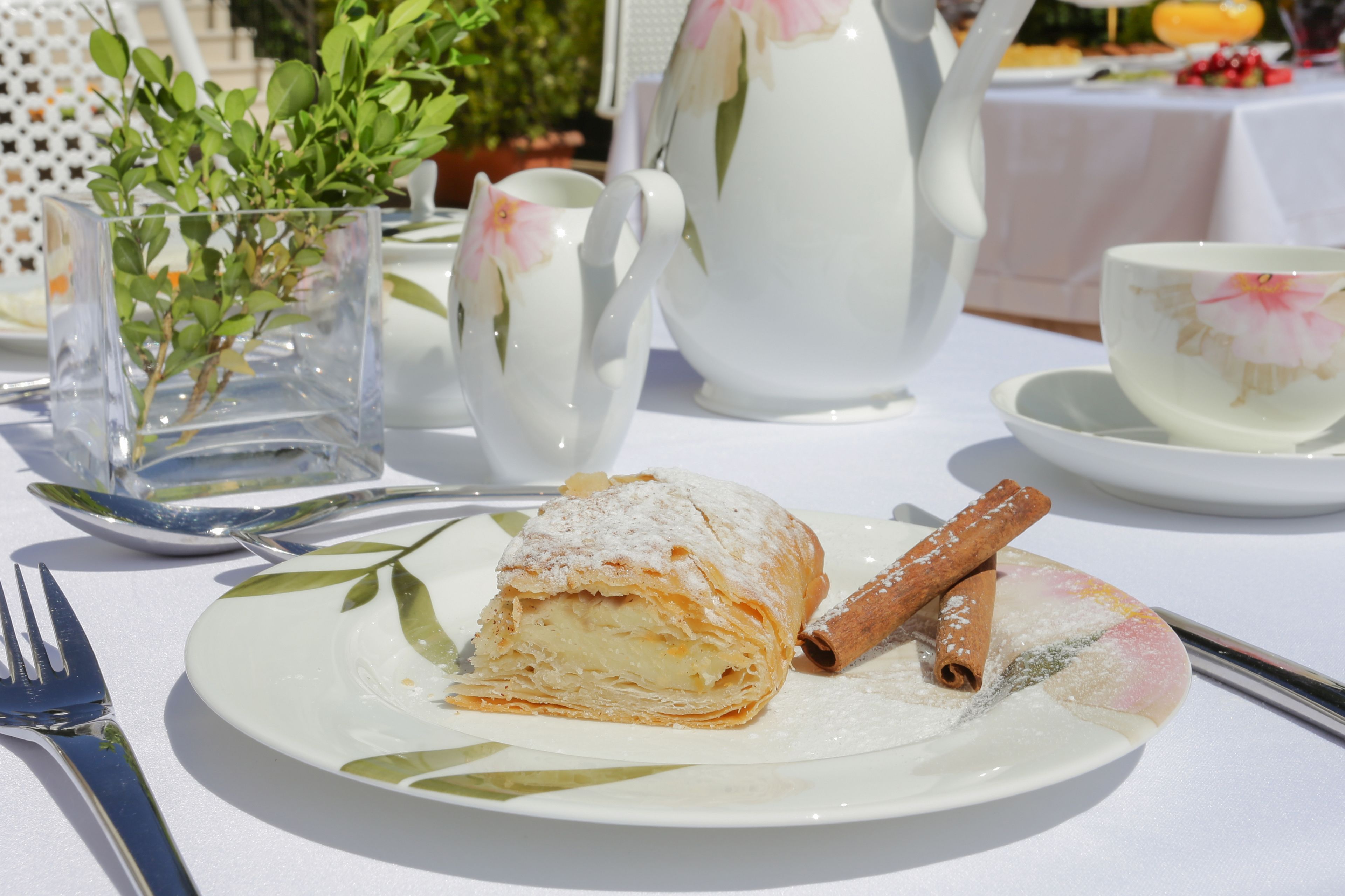 A table set with a plate of pastry, a teapot, a cup, and a vase with green plants.